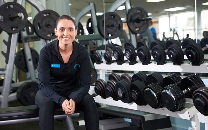 A photo of a gym staff member sitting on a bench next to a rack of free weights